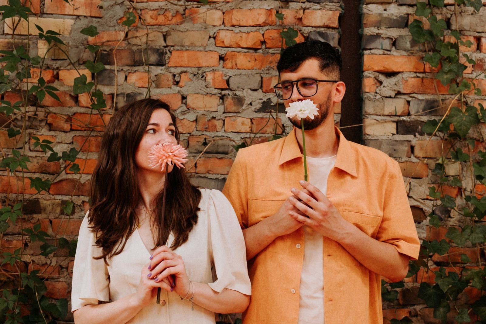 Romantyczna sesja z parą trzymającą różową dalię i białą przed rustykalną ceglaną ścianą.